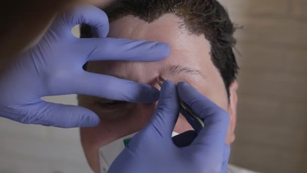 Procedimiento cosmético para el cuidado de las cejas durante el virus. Una mujer con una máscara y guantes arranca unas pinzas con una ceja a un cliente enmascarado durante el virus — Vídeos de Stock