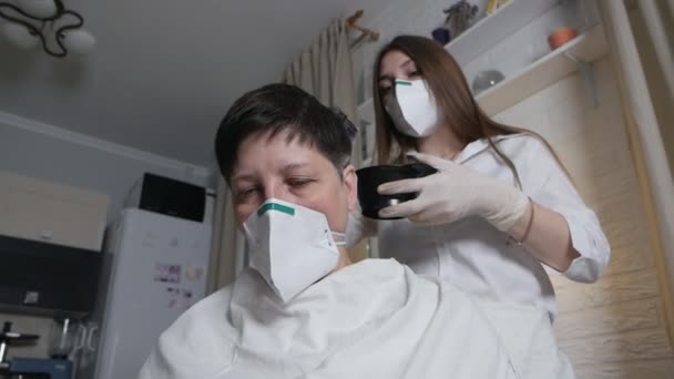 Μια νεαρή γυναίκα με ιατρική μάσκα βάφει τα μαλλιά της μητέρας της στο σπίτι κατά τη διάρκεια της επιδημίας του ιού. Αυτοφροντίδα στο σπίτι — Αρχείο Βίντεο
