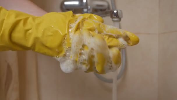 En kvinna tvättar händerna i gula gummihandskar i handfatet under kranen med vatten. Hemmafru tvättar tvål med skyddshandskar efter städning. Begreppet städning och renlighet — Stockvideo