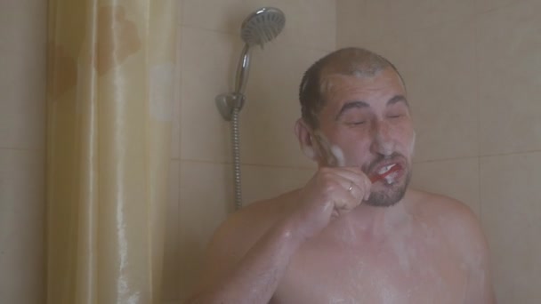 Un uomo in piedi in bagno si lava i denti e si spruzza con acqua dalla doccia — Video Stock