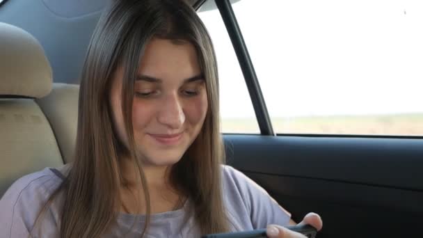 Glimlachend meisje zittend op de achterbank van een auto tijdens het reizen met behulp van een mobiele telefoon kijken naar een film — Stockvideo
