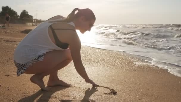 La chica se dibuja en la arena cerca de las olas del mar al atardecer. Disparos en la playa — Vídeo de stock