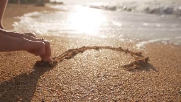 La chica se dibuja en la arena cerca de las olas del mar al atardecer. Disparos en la playa — Vídeos de Stock