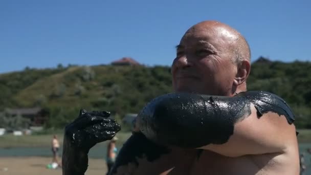 En gammal man applicerar svart helande lera på sin kropp på en saltvattensjö. Svart lera, lera är användbart för personer med hälsa och hudproblem. Koppla av på spaanläggningen — Stockvideo