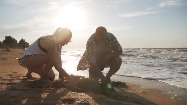En una tarde de verano, un hombre se arrodilla y cava un agujero con las manos en la playa junto al mar. Familia feliz: padre y su hija adulta están construyendo un castillo de arena en la playa del mar — Vídeo de stock