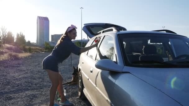 De vrouw is bij de auto. Jonge mooie vrouw met een mooie figuur veegt het glas van de auto bij zonsondergang — Stockvideo