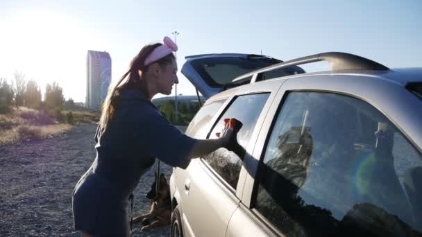 Hermosa mujer joven limpia el vidrio en la puerta del coche con un trapo en la calle al atardecer. — Vídeo de stock