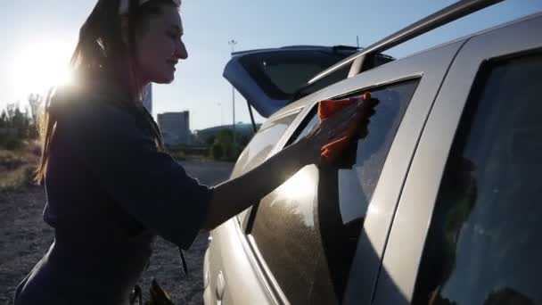 Hermosa mujer joven limpia el vidrio en la puerta del coche con un trapo en la calle al atardecer — Vídeo de stock