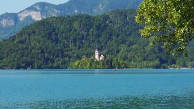 Arka planda dağlarla çevrili bir kilise adası ve Slovenya sularındaki yansımalarla dolu panoramik göl manzarası