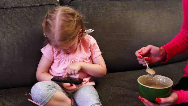 Una bambina guarda il suo smartphone mentre sua madre le nutre un cucchiaio — Video Stock