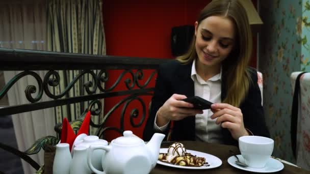 Una joven con chaqueta y blusa en un café toma una foto del postre y selfie — Vídeos de Stock