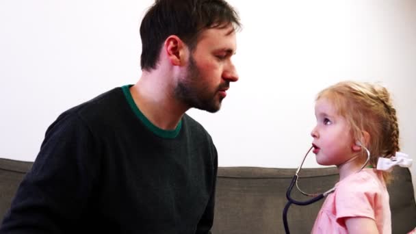 Ein Vater und seine süße Tochter spielen in Arzt. ein Baby lauscht mit seinem Stethoskop der Lungenarbeit eines Vaters. — Stockvideo