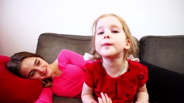 Una niña dice y mira en cámara. Una madre acostada en el sofá . — Vídeos de Stock