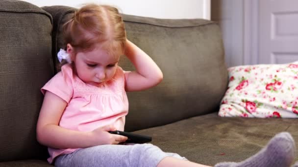 Маленькая девочка играет со смартфоном и смотрит в камеру — стоковое видео