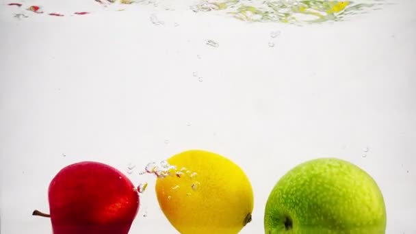 La manzana, el limón y el naranja caen en el agua con burbujas en movimiento lento. Frutos aislados de un fondo blanco. — Vídeos de Stock