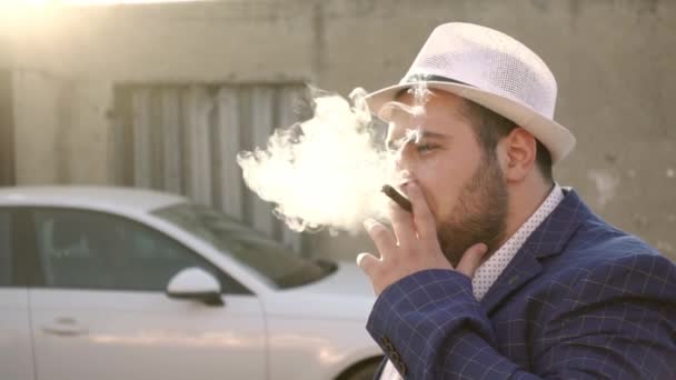 Brodaty mężczyzna w kapeluszu i garnitur pali cygaro na parkingu w zwolnionym tempie, widok z boku. — Wideo stockowe
