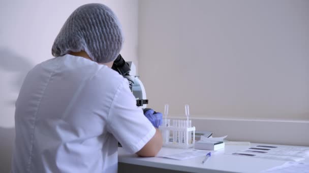 Assistente de laboratório senta-se em seu local de trabalho com um microscópio de laboratório — Vídeo de Stock
