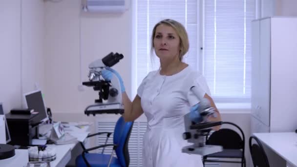 Hermosa rubia en bata médica baila en laboratorio con microscopios — Vídeos de Stock