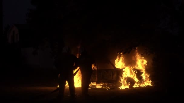 Deux silhouettes pompiers se tiennent devant une voiture en feu au ralenti — Video