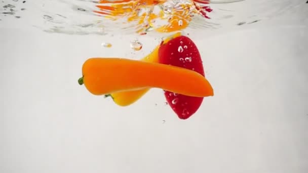 Três pimentas frescas multicoloridas caem na água no centro com salpicos e bolhas — Vídeo de Stock