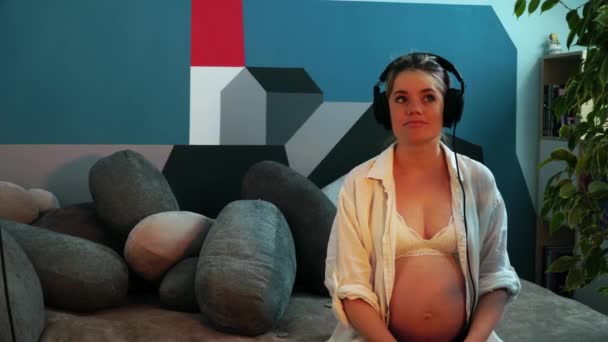 Pregnant woman sitting in headphones and puts it to her bare belly — Stock Video