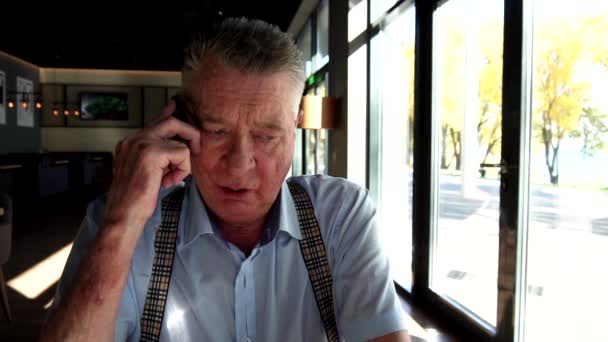 El viejo está sentado cerca de la ventana en el restaurante y hablando por teléfono con tristeza — Vídeo de stock