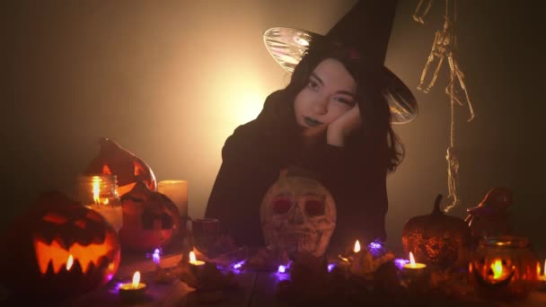 Jovem bruxa no manto e chapéu preto está sentado entediado com o rosto na mão — Vídeo de Stock