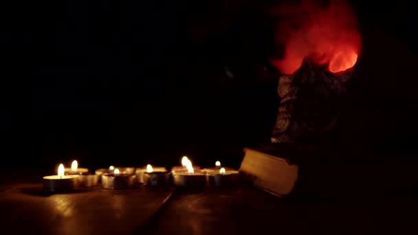 Czaszka człowieka na książce z świecącymi czerwonymi oczami, z których wydobywa się dym na tle świec — Wideo stockowe