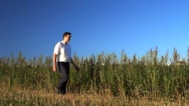 Genç beyaz bir adam yaz tarlasında yürür. Yavaş çekimde çimenlere dokunur.