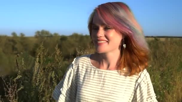 Młoda uśmiechnięta dama z wielobarwnymi włosami spaceruje po polu i dotyka trawy w slowmo — Wideo stockowe