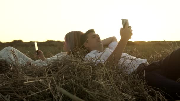 Slowmo de casal caucasiano que está deitado no palheiro no campo ao pôr do sol e observando seus celulares — Vídeo de Stock