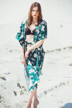 Yeşil mayo beyaz kum plaj üzerinde poz çekici ince kız. Genç kadın tatil kumlu okyanus sahilinde bikin dayanmaktadır. Kavram seyahat, mayo