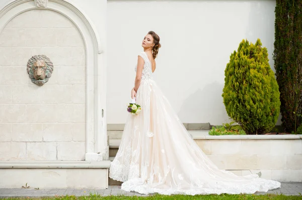 結婚式の前に白い家の近くを歩く光の長い白いドレスで結婚式の花束を持つ若いエレガントな女性 — ストック写真