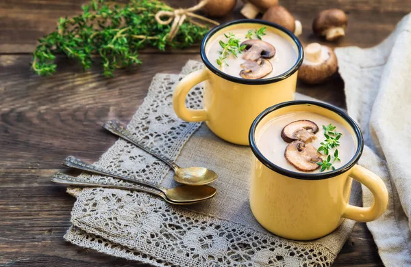 Creamy mushroom soup with champignons in mugs on rustic wooden background.