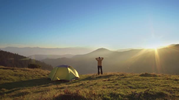 Мужчина стоит рядом с палаткой в горах — стоковое видео