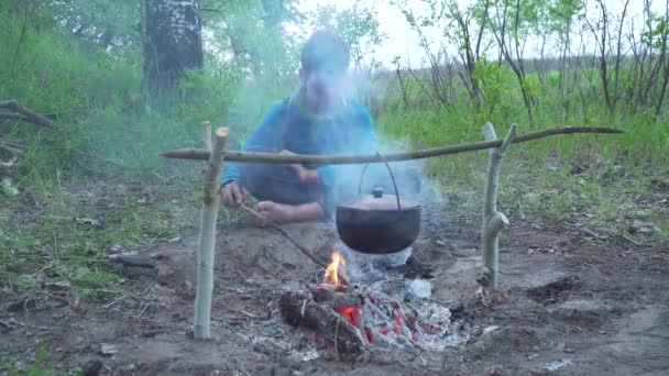 Un niño se sienta junto a un fuego en el bosque — Vídeo de stock