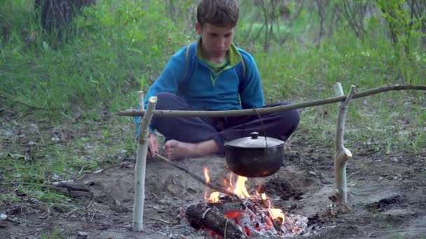 Een jongen zit bij een vuur in het bos — Stockvideo