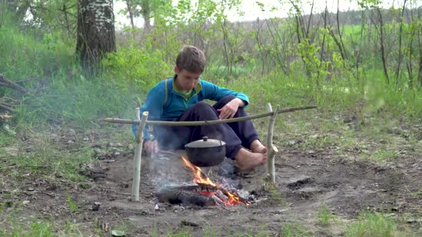 En pojke sitter vid en eld i skogen — Stockvideo