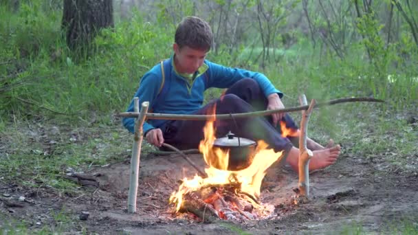 Een jongen zit bij een vuur in het bos — Stockvideo
