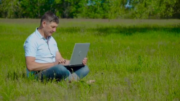 Uomo d'affari lavora dietro un computer portatile seduto sull'erba — Video Stock