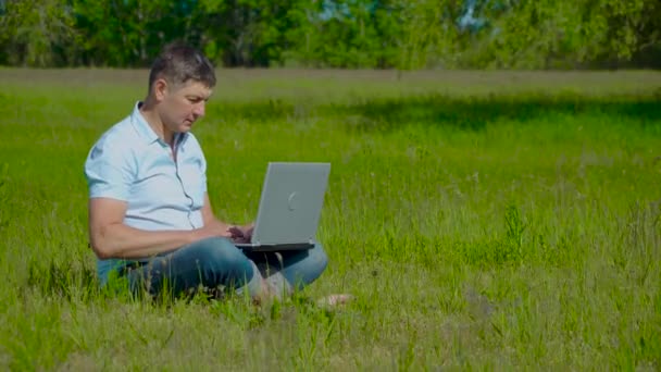 Бизнесмен работает за ноутбуком, сидя на траве — стоковое видео