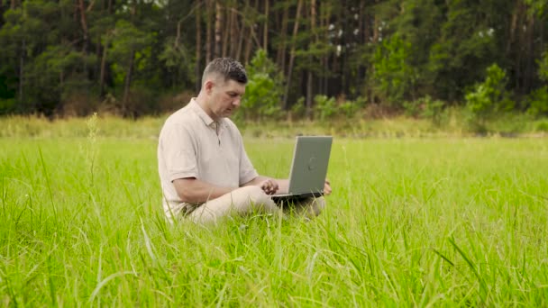 Serbest İşçi, çimenlikteki çimlerde otururken uzaktan kumanda için dizüstü bilgisayar kullanıyor. — Stok video
