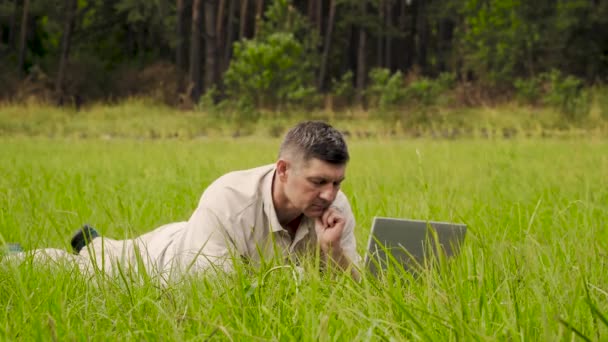 Бизнесмен работает за ноутбуком, лежащим на траве — стоковое видео