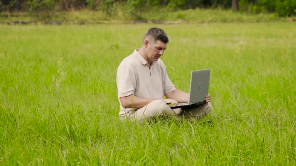 Freelancer использует ноутбук для удаленной работы, сидя на газоне в траве — стоковое видео