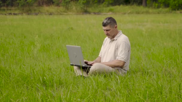 Freelancer использует ноутбук для удаленной работы, сидя на газоне в траве — стоковое видео