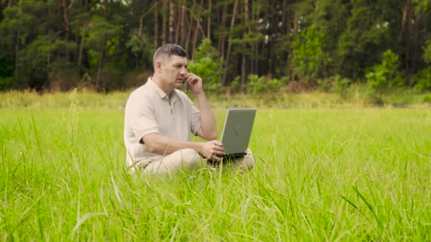 Facet z laptopem rozmawia przez telefon na zielonym trawniku. — Wideo stockowe