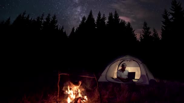 Чоловік працює за ноутбуком у наметі біля багаття вночі — стокове відео