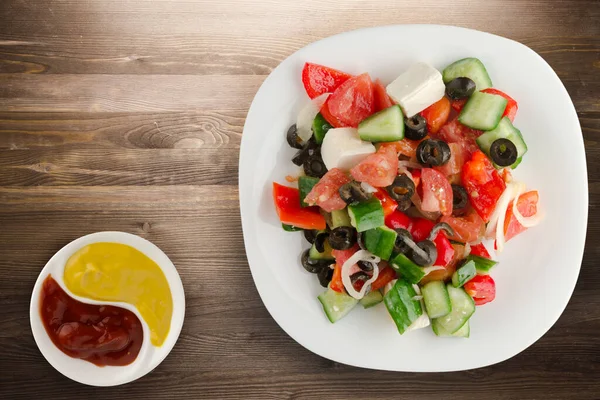 Kahverengi ahşap arka planda Yunan salatası. Beyaz tabakta Yunan salatası ve sos manzarası. sağlıklı yiyecekler