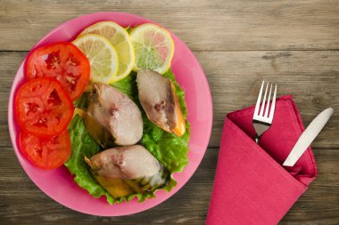 Çatal ve bıçak manzaralı pembe bir tabakta tütsülenmiş uskumru. Kahverengi ahşap arka planda limonlu uskumru ve domates.