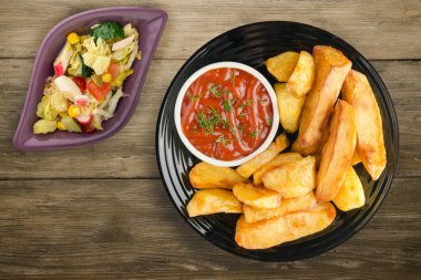 Kahverengi ahşap arka planda ketçaplı patates kızartması. Siyah tabakta patates kızartması ve vejetaryen salatası.